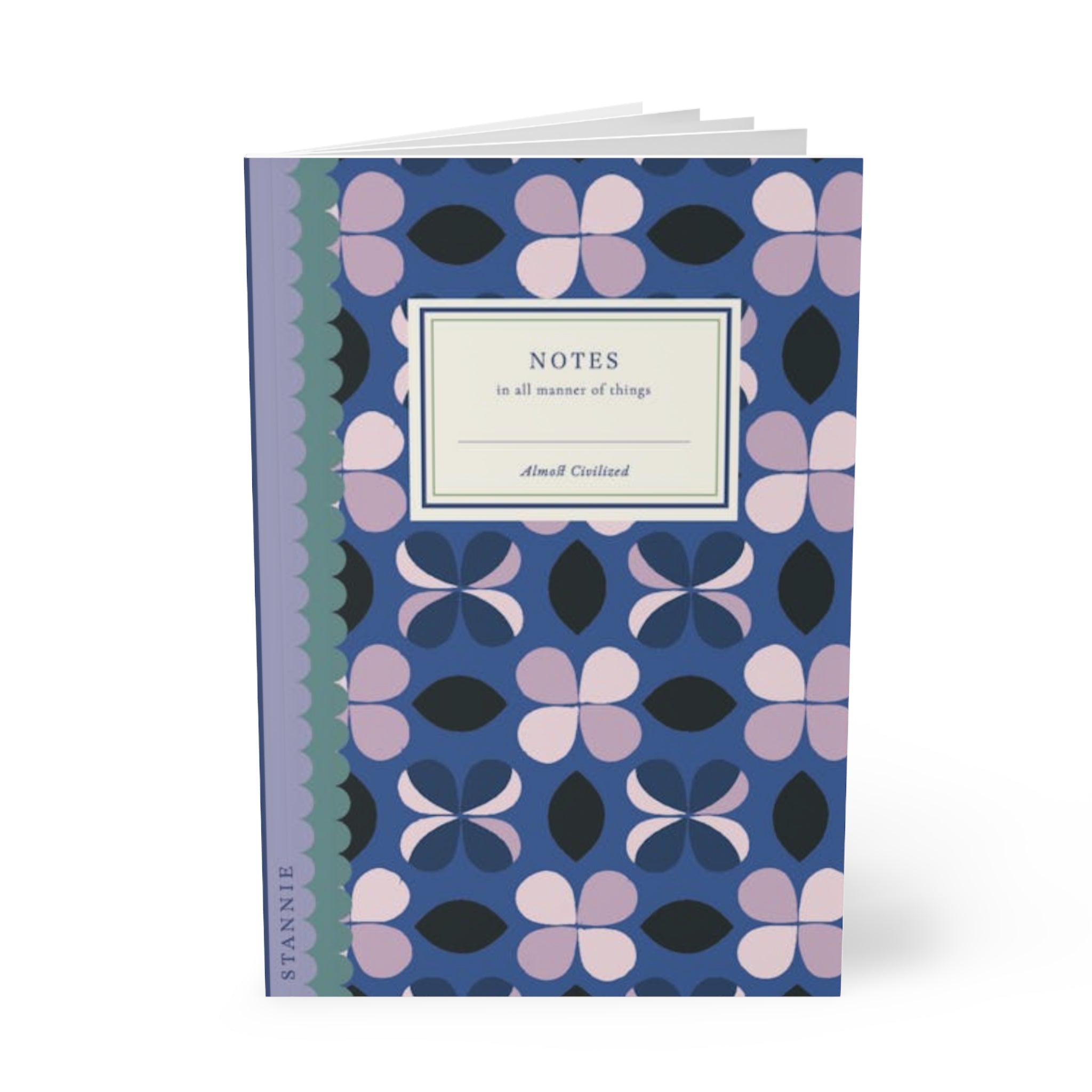 A blue geo Softcover Notebook, A5 by Printify with a laminated cover adorned in pink and black floral designs, titled "NOTES in all manner of things." The author's name, "STANNIE," appears on the spine. Inside, you'll find high-quality 90gsm lined paper perfect for all your notes.