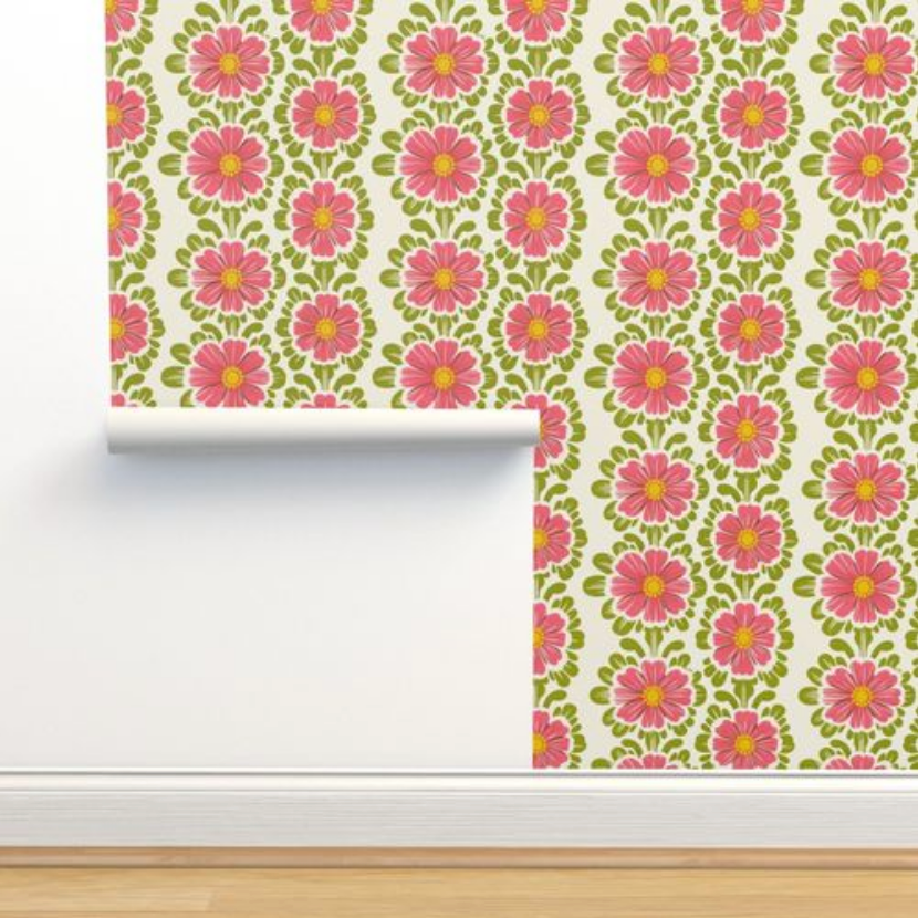 Roll of Pink Pop Flowers Wallpaper by Stannie & Lloyd with pink and yellow flowers against a green background, partially unrolled against a white wall above a wooden floor.